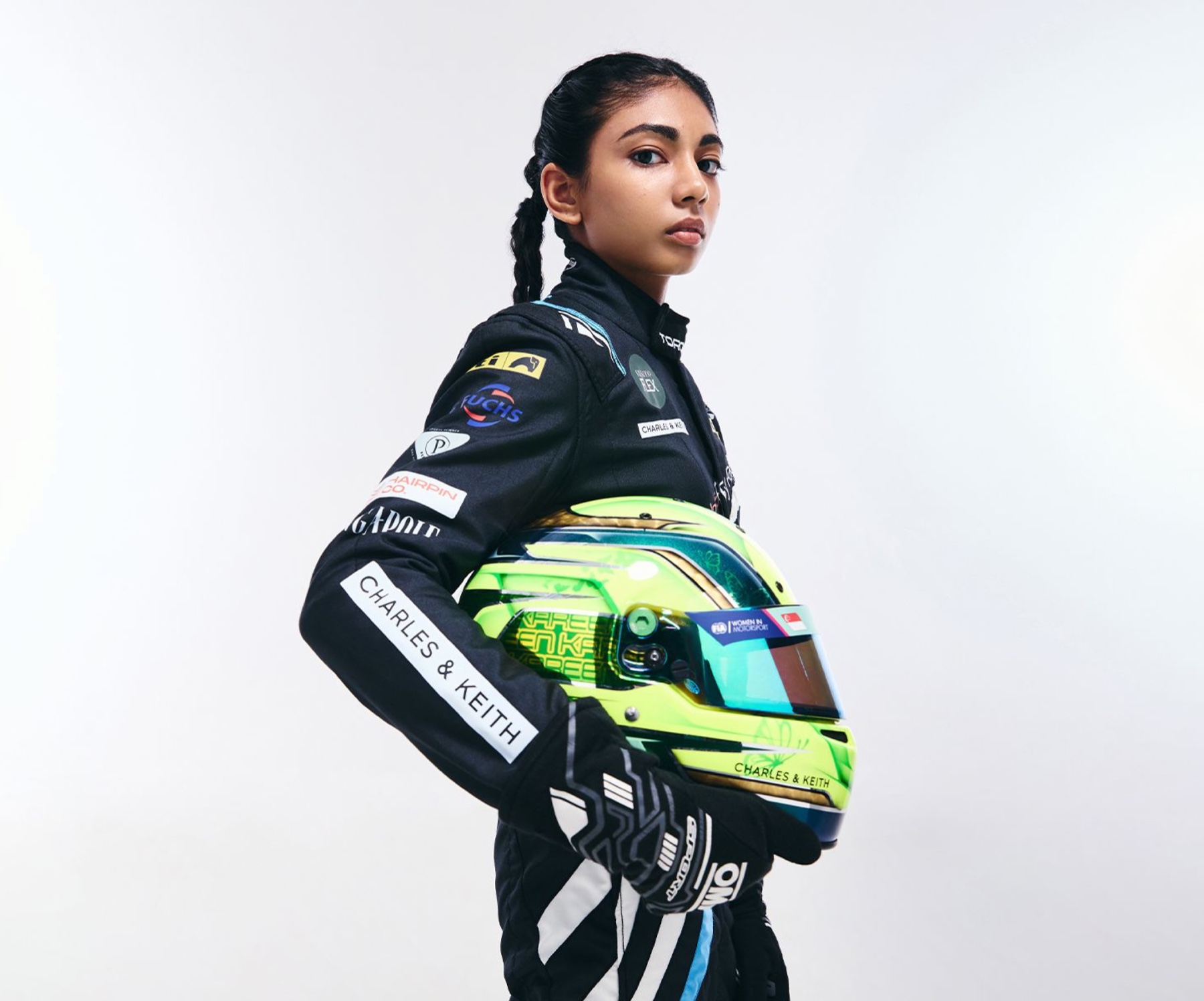 Kareen Kaur in a black race suit holding a green and yellow helmet against a light studio backdrop.