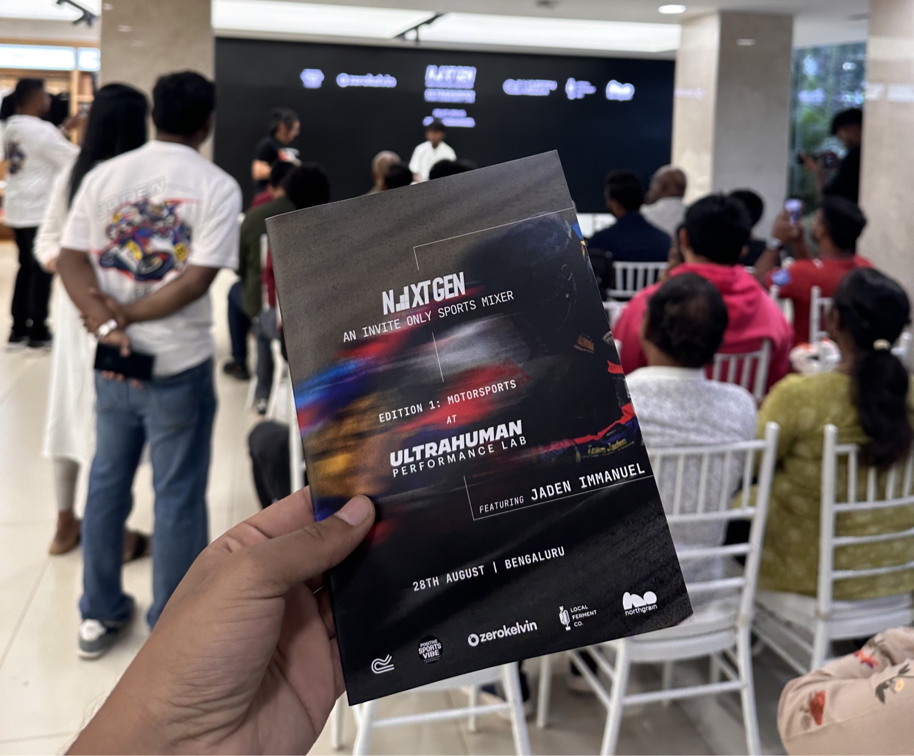 A hand holding a NextGen event flyer in front of a seated audience.